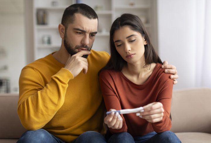 Infertility Problems. Portrait Of Upset Young Couple Looking At Negative Pregnancy Test While Sitting Together On Couch At Home, Caring Husband Embracing Depressed Wife, Free Space